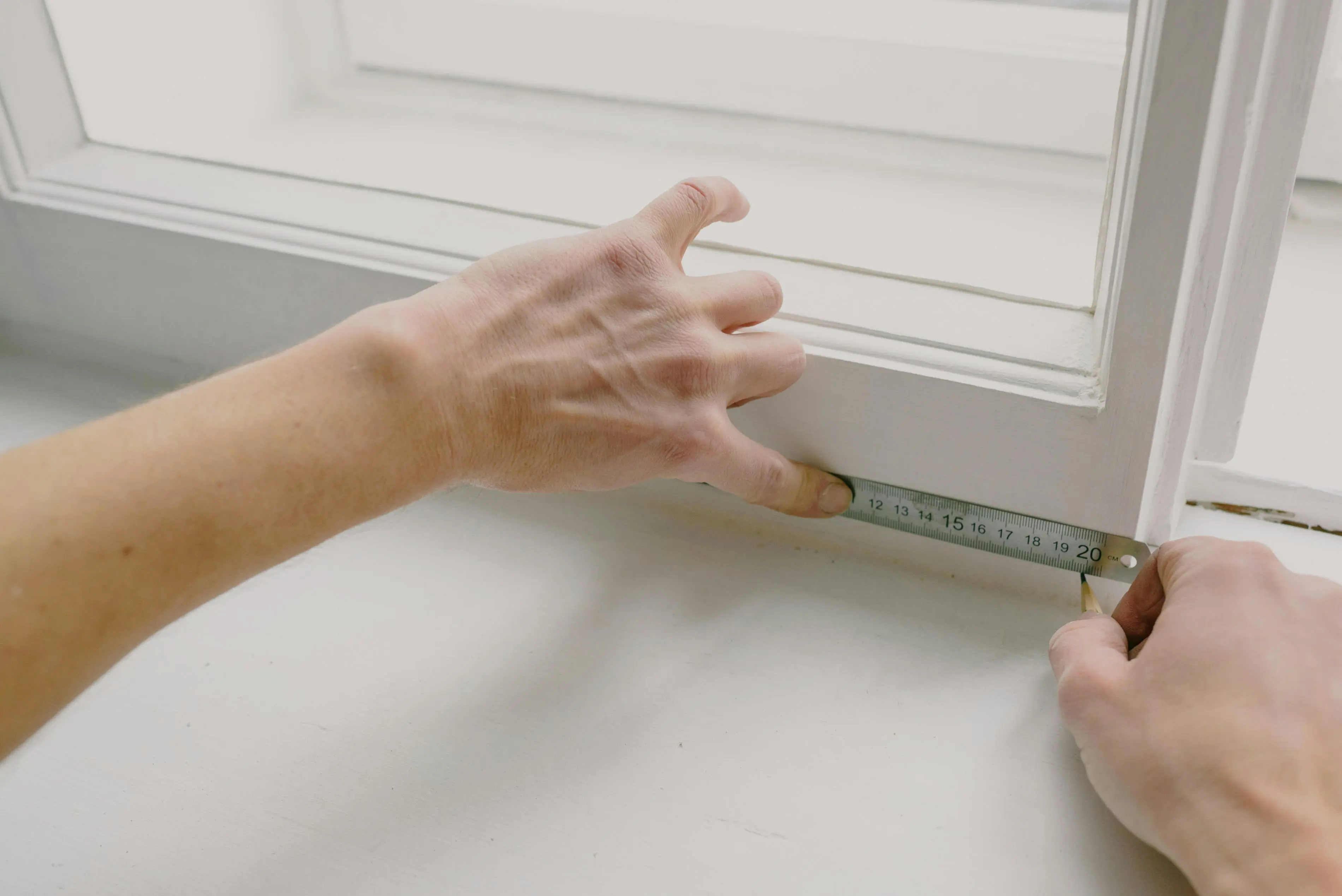 Gros plan sur des mains mesurant l'embrasure d'une fenêtre blanche à l'aide d'un mètre pliant, en atelier.