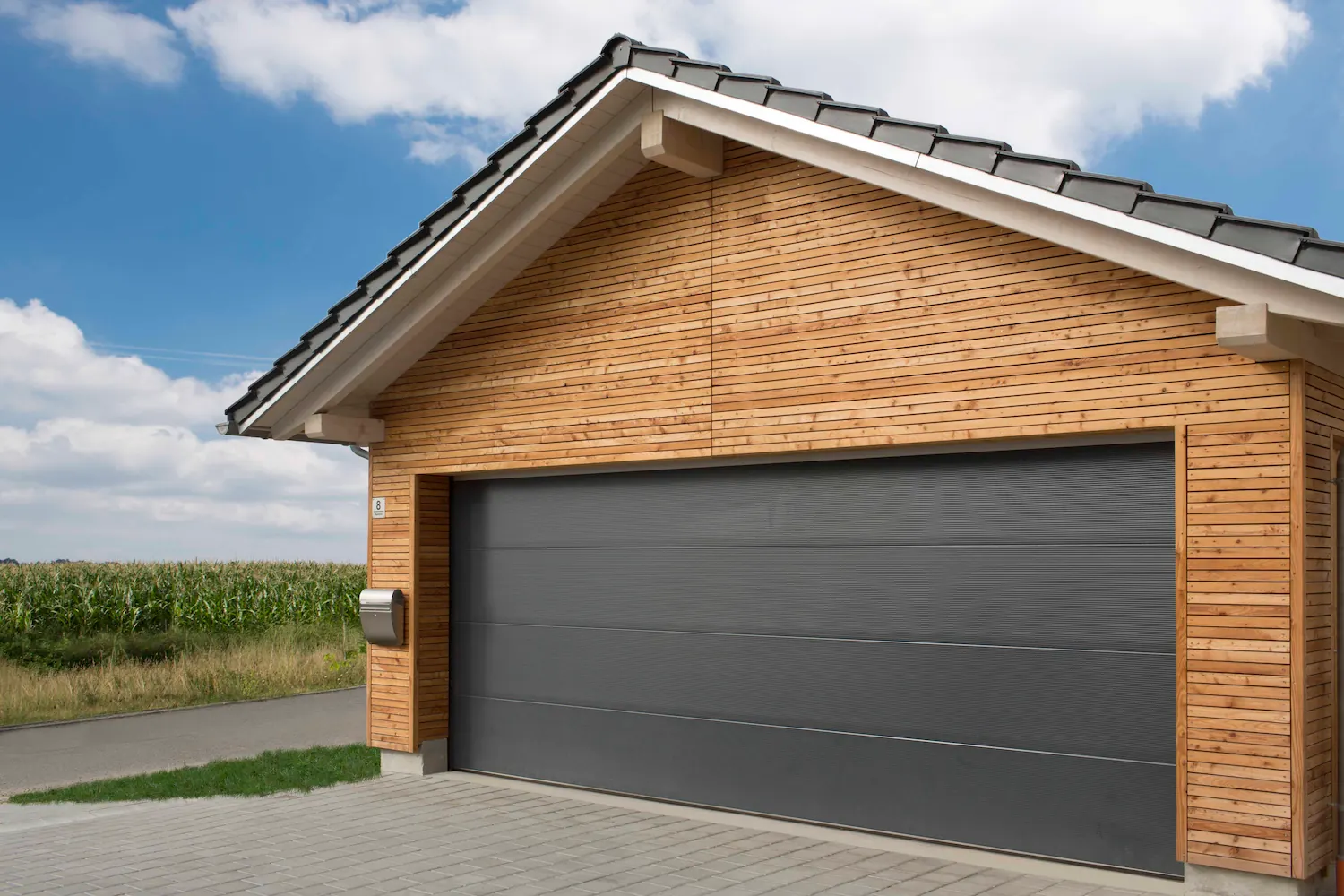 Porte de garage sectionnelle Normstahl imitation bois gris anthracite, intégrée à un pignon à bardage bois clair sous un toit à tuiles, en bordure d'un champ.