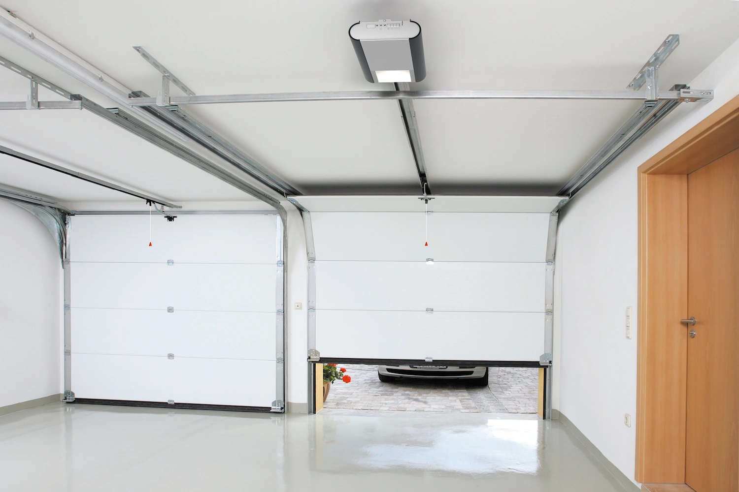 Vue intérieure d'un garage double avec deux portes sectionnelles blanches, moteur d'ouverture automatique Normstahl fixé au plafond et porte de service en bois.