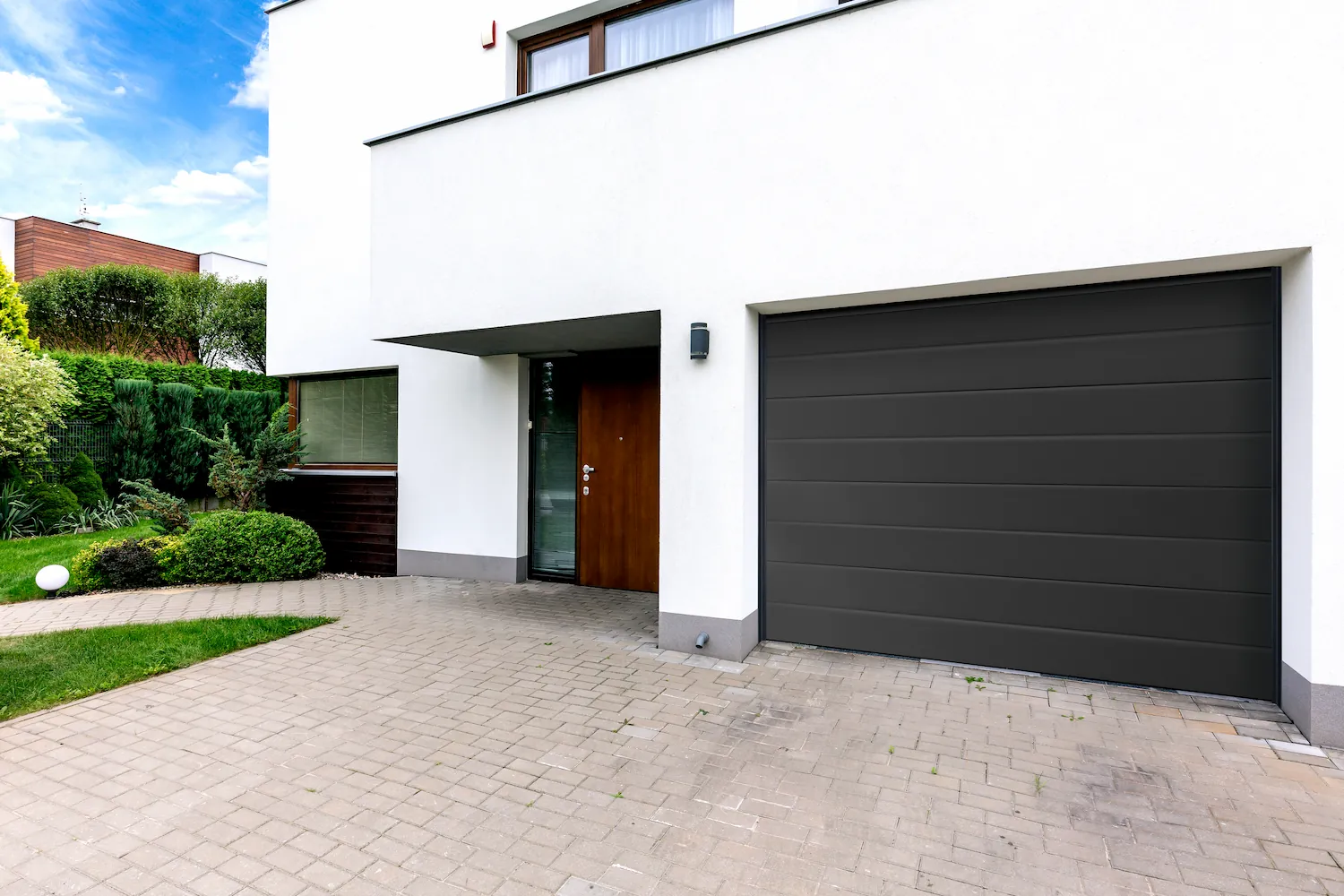 Porte de garage sectionnelle Normstahl gris anthracite à lames horizontales, intégrée à une villa moderne blanche avec porte d'entrée en bois attenante.