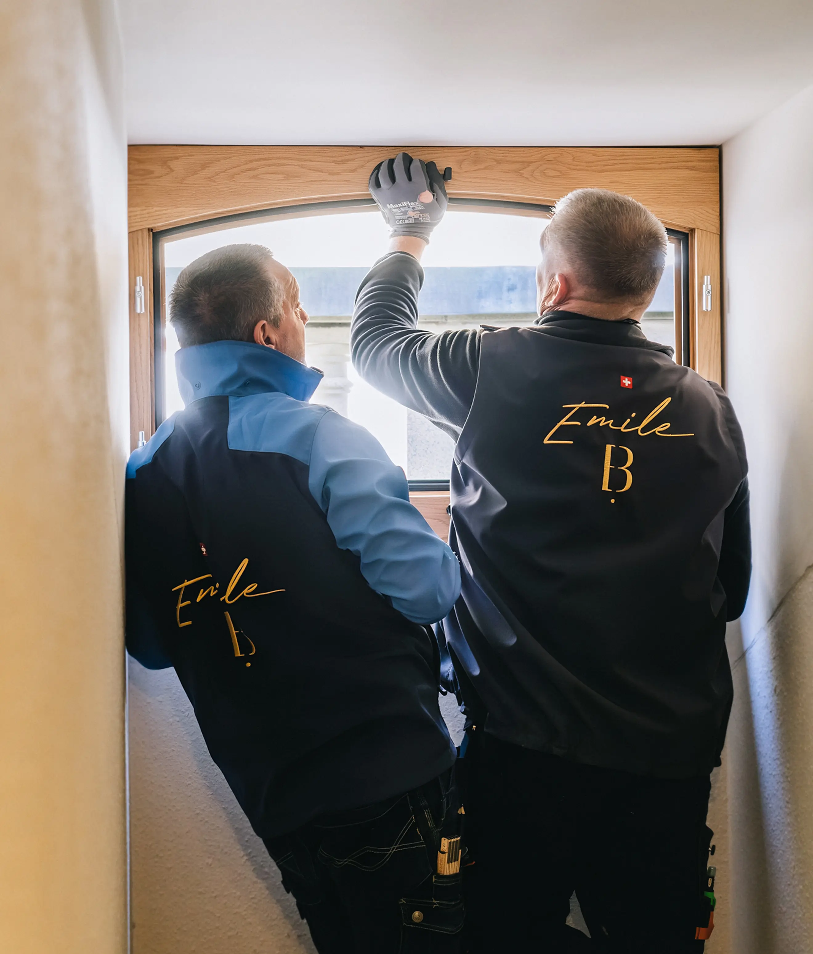 Deux artisans menuisiers de dos, en vestes de travail brodées, installant une fenêtre cintrée en bois de chêne dans une embrasure de mur.