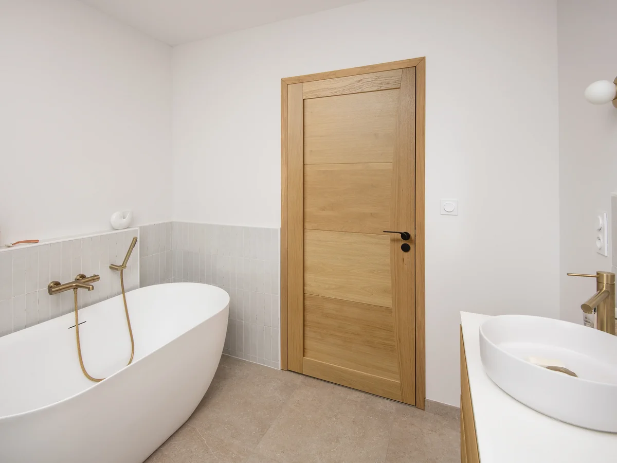 Porte de salle de bain en bois clair à panneaux horizontaux avec poignée noire, dans une salle de bain avec baignoire îlot blanche et robinetterie dorée.