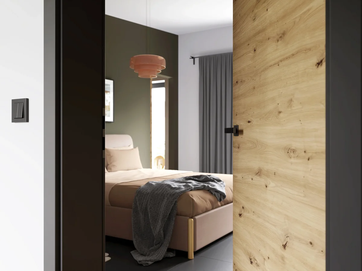 Porte intérieure en bois clair entrebâillée sur une chambre aux tons taupe, avec suspension cuivrée et lit habillé de plaid gris.