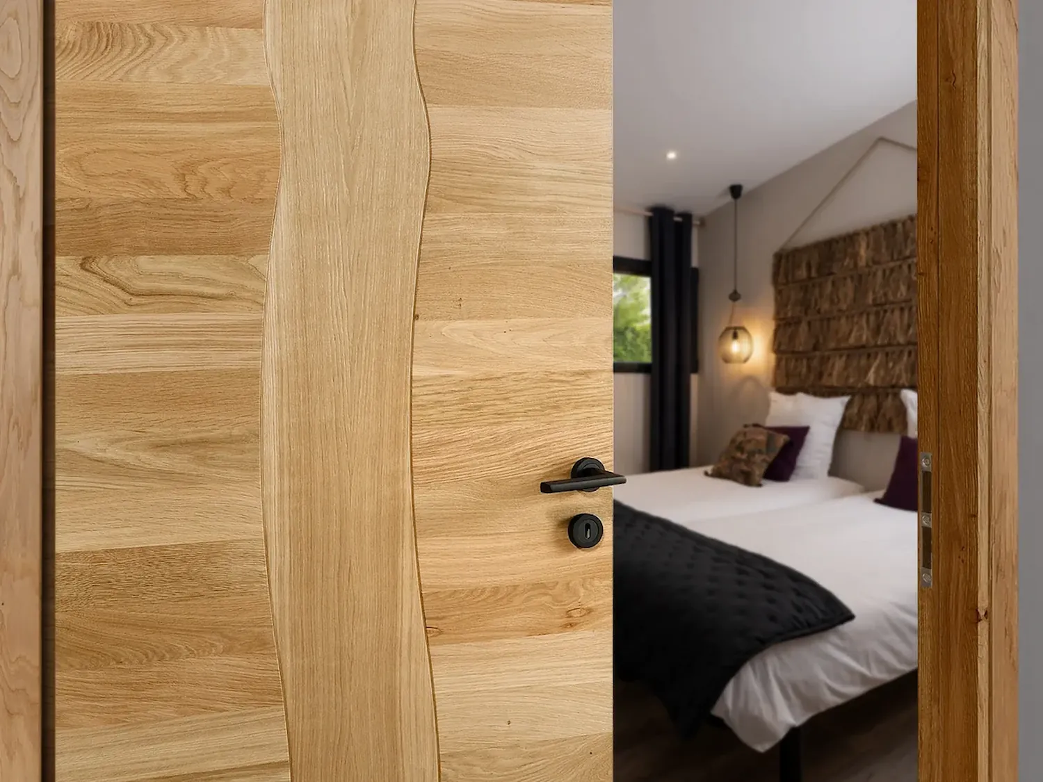Porte intérieure en bois chêne clair entrebâillée avec poignée noire, ouvrant sur une chambre à coucher au linge bordeaux et blanc.