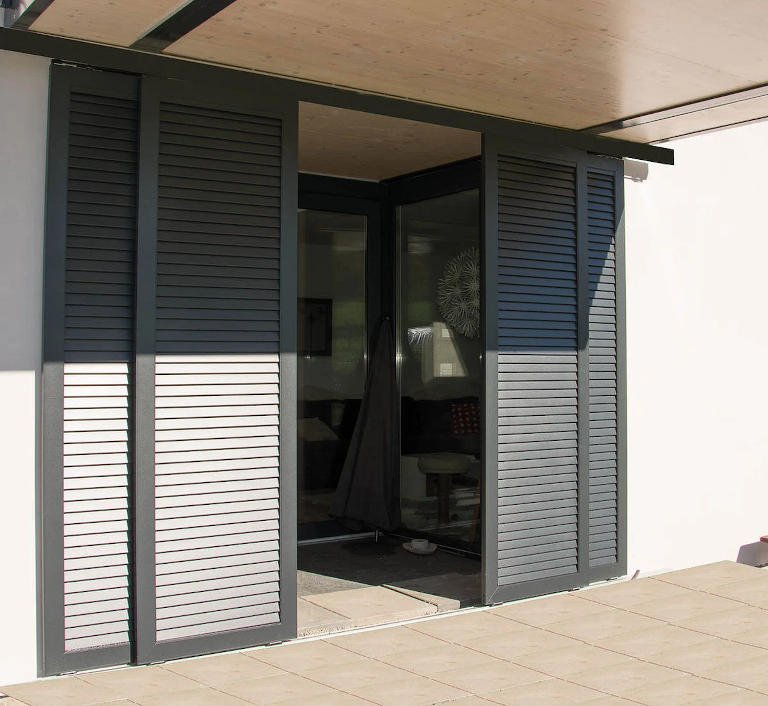 Angle de terrasse couverte avec volets coulissants en aluminium gris anthracite à lames persiennées, partiellement ouverts sur un salon intérieur.