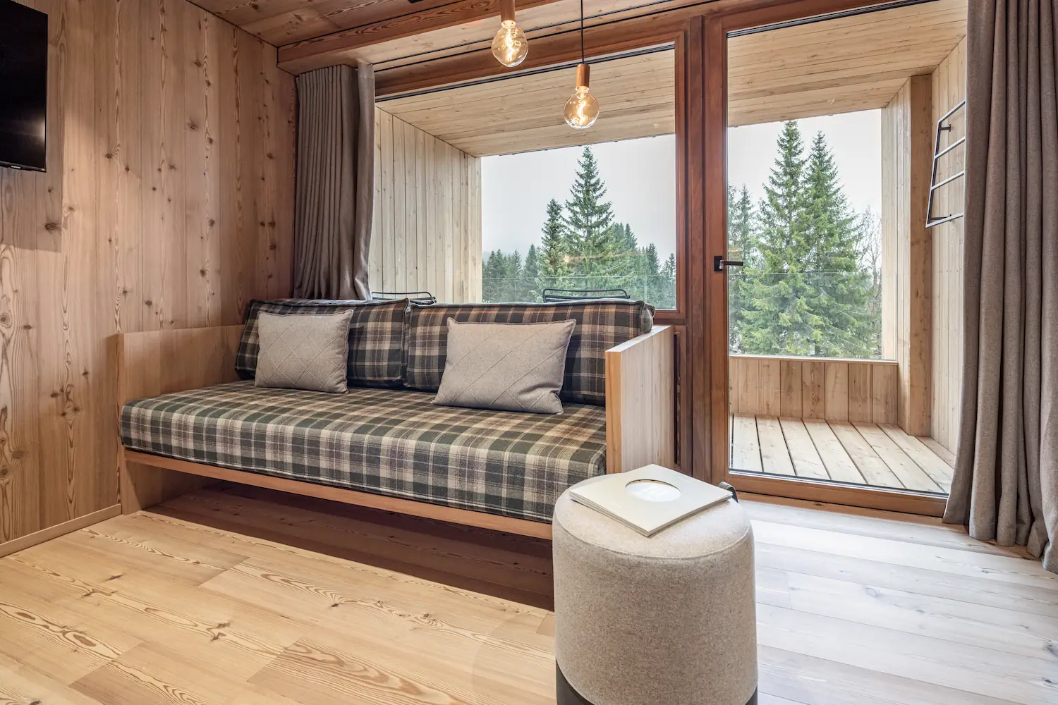 Intérieur chalet en bois avec banquette à coussins à carreaux et porte-fenêtre coulissante en bois ouvrant sur un balcon avec vue sur des sapins enneigés.