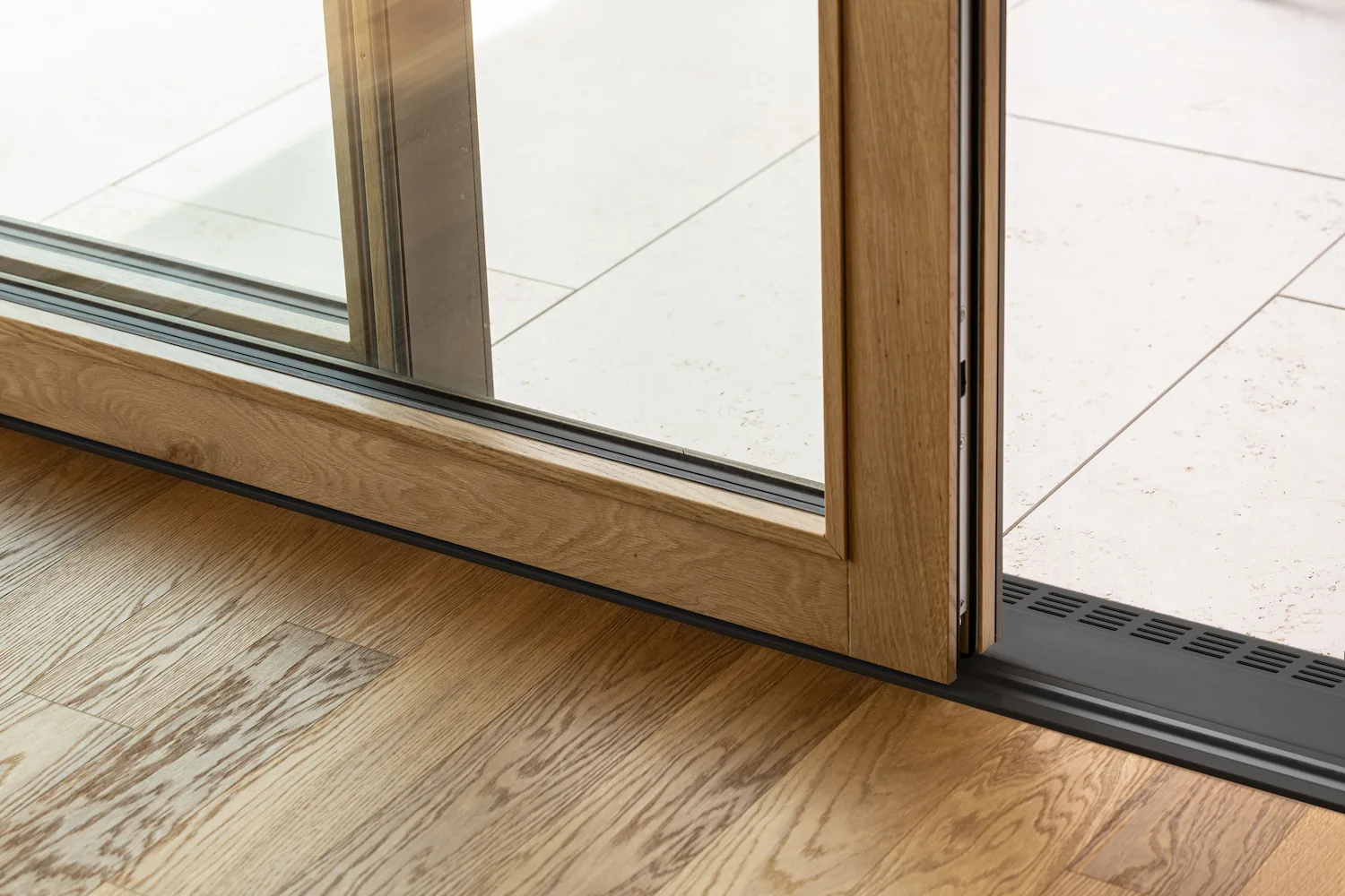 Détail du seuil d'une baie vitrée coulissante à cadre bois, entre un parquet clair intérieur et une terrasse carrelée extérieure.