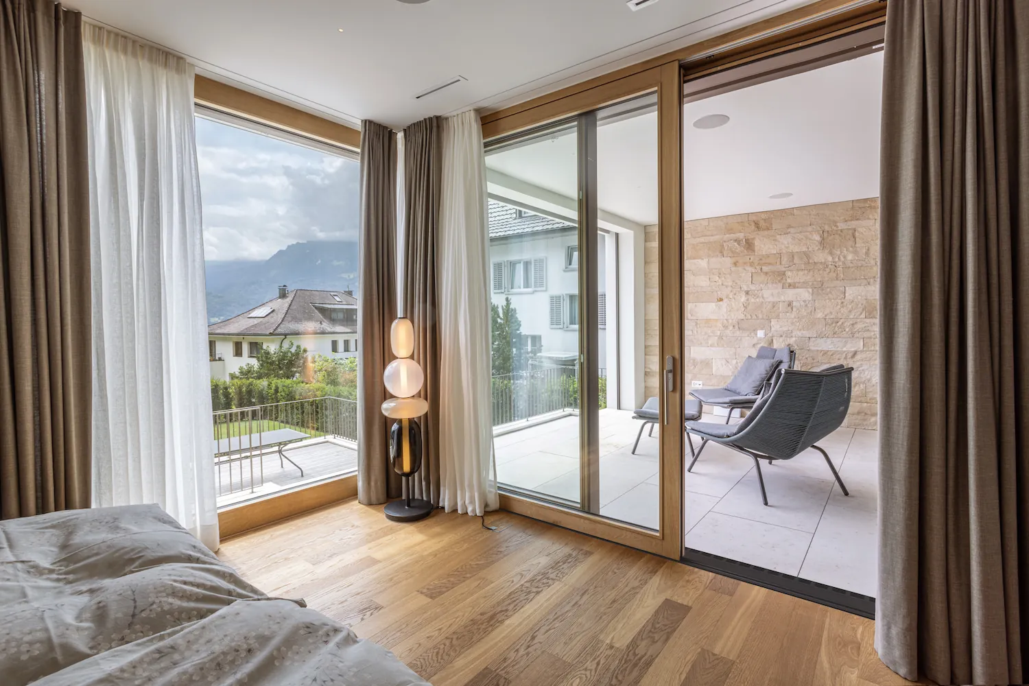 Chambre lumineuse avec baie vitrée coulissante bois-aluminium ouvrant sur une terrasse avec vue montagne, rideaux voilage et parquet clair.