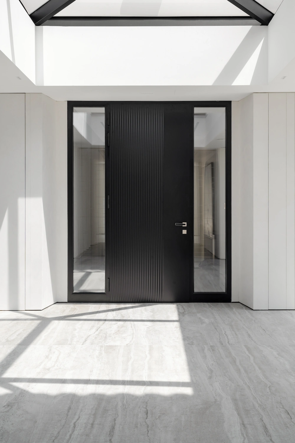 Porte d'entrée pivotante noire à cannelures verticales sous une verrière triangulaire, encadrée de fixes vitrés, dans un hall lumineux au sol en travertin.