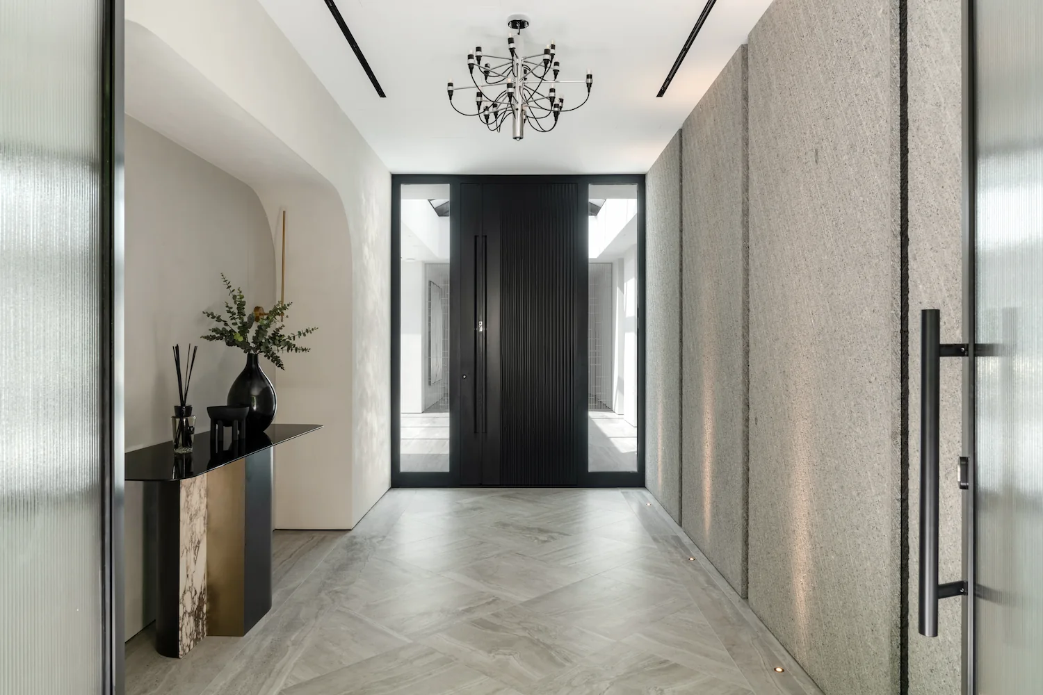 Porte d'entrée pivotante noire à cannelures verticales, encadrée de deux fixes vitrés, dans un hall d'entrée en marbre gris avec lustre design.