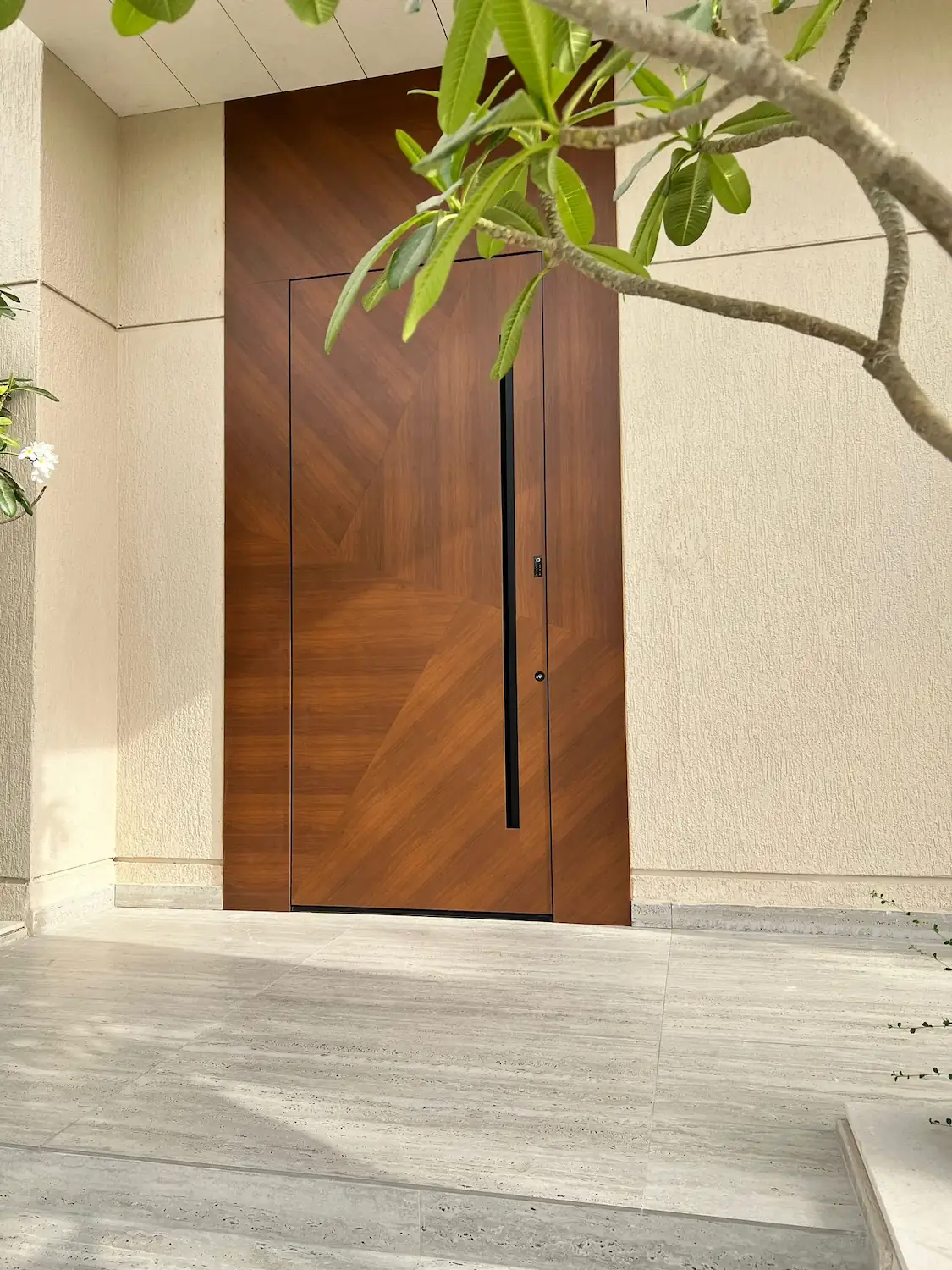 Porte d'entrée pivotante en bois brun à motif chevron, avec longue poignée verticale noire, sur une façade beige ombragée par un frangipanier.