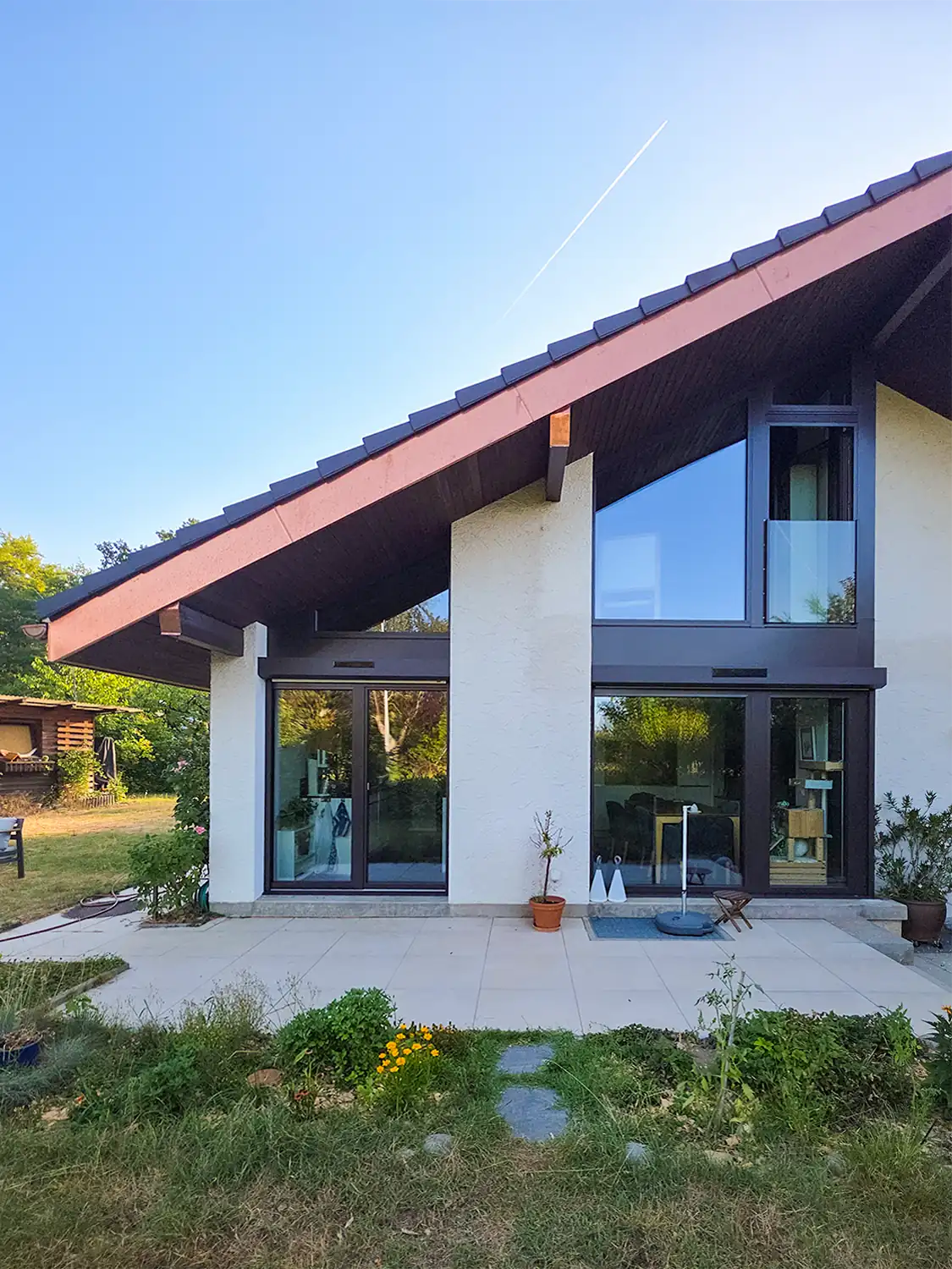 Extension contemporaine d'une maison à toit à un pan et grandes baies vitrées à cadres anthracite, terrasse dallée, photographiée en fin de journée.