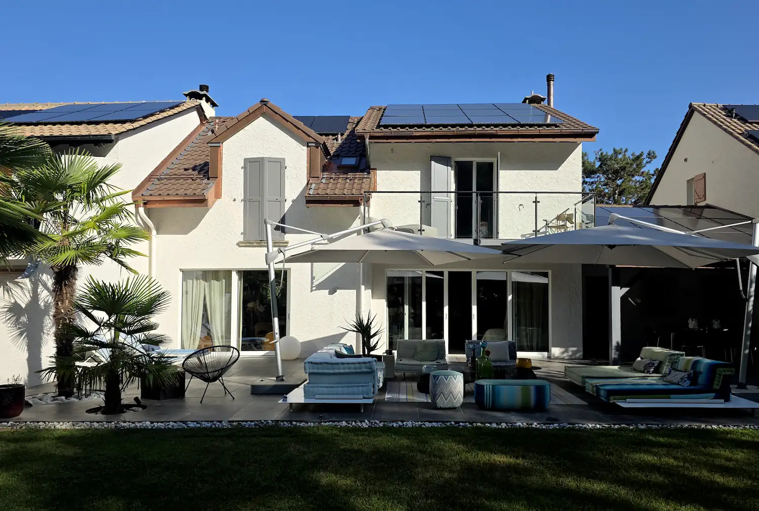Façade arrière d'une villa équipée de panneaux solaires en toiture, avec terrasse aménagée d'une pergola à voiles blanches et mobilier de jardin.