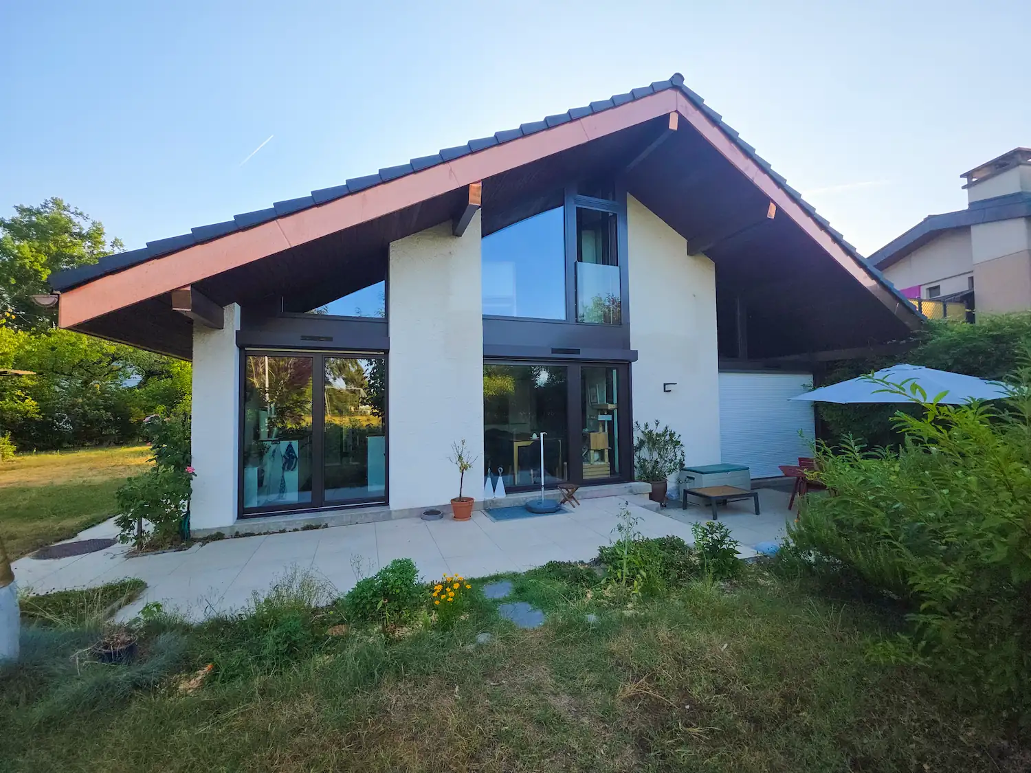 Extension contemporaine d'une maison à ossature bois et toit à un pan asymétrique, grandes baies vitrées et terrasse aménagée, photographiée au coucher du soleil.