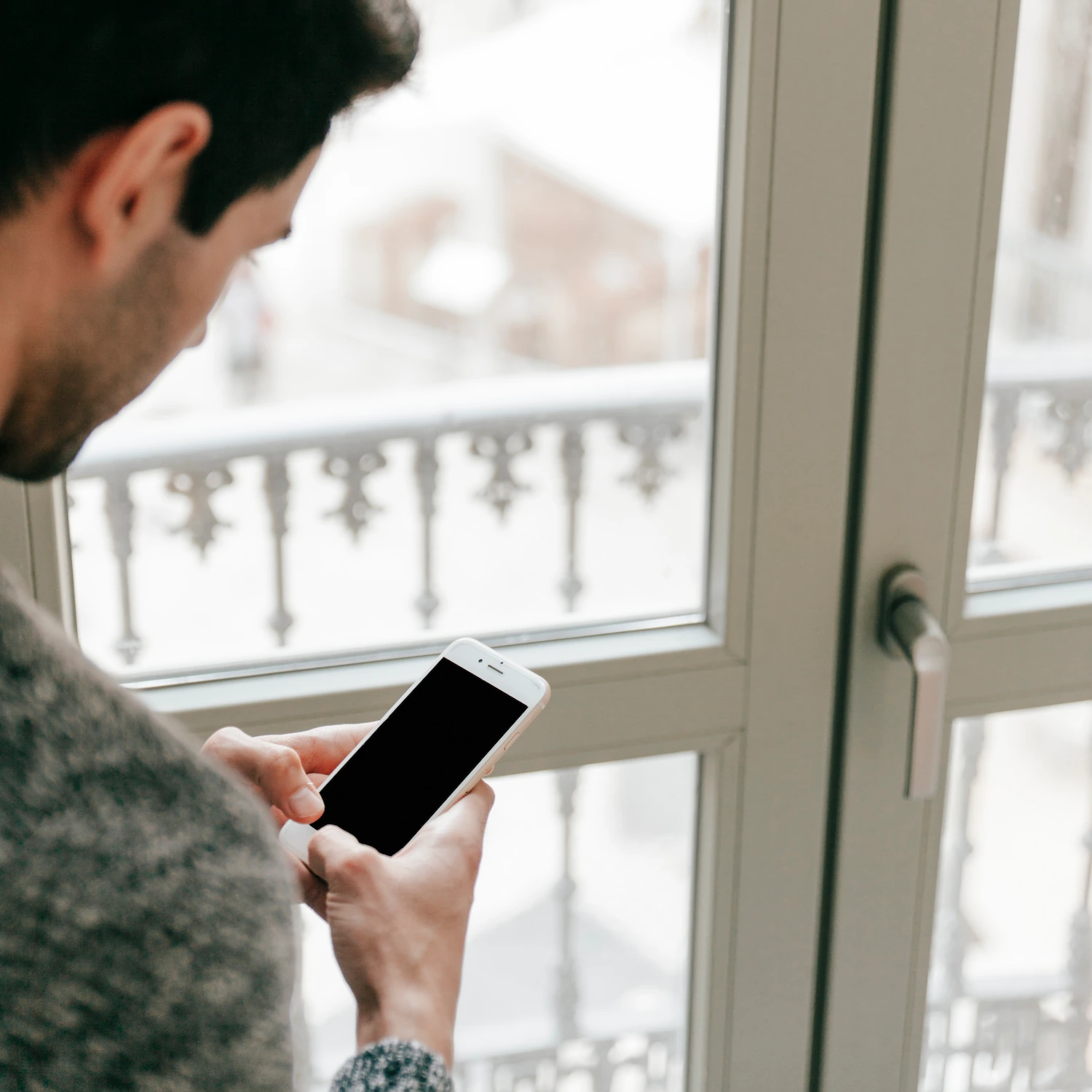Homme de dos consultant son smartphone près d'une porte-fenêtre à balcon parisien, vue enneigée à l'extérieur.