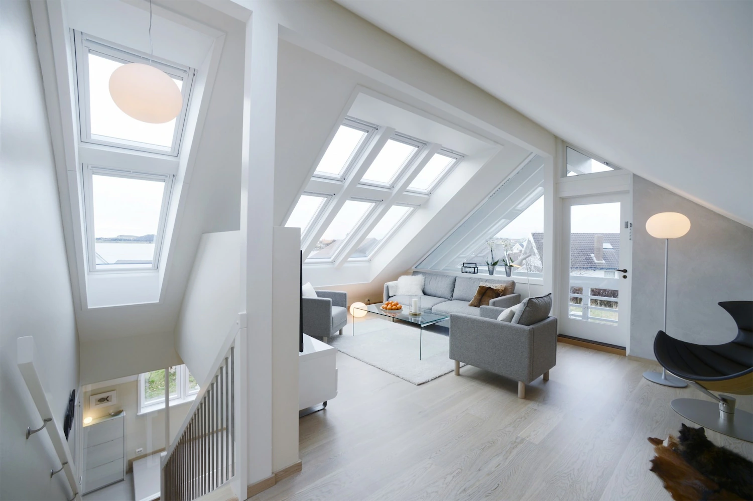 Salon mansardé lumineux aménagé sous plusieurs fenêtres de toit Velux, avec canapés gris, coin télévision et vue sur la mer.