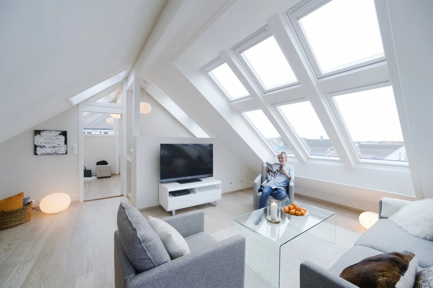 Salon aménagé sous combles éclairé par plusieurs fenêtres de toit Velux inclinées, avec canapés gris et coin télévision.