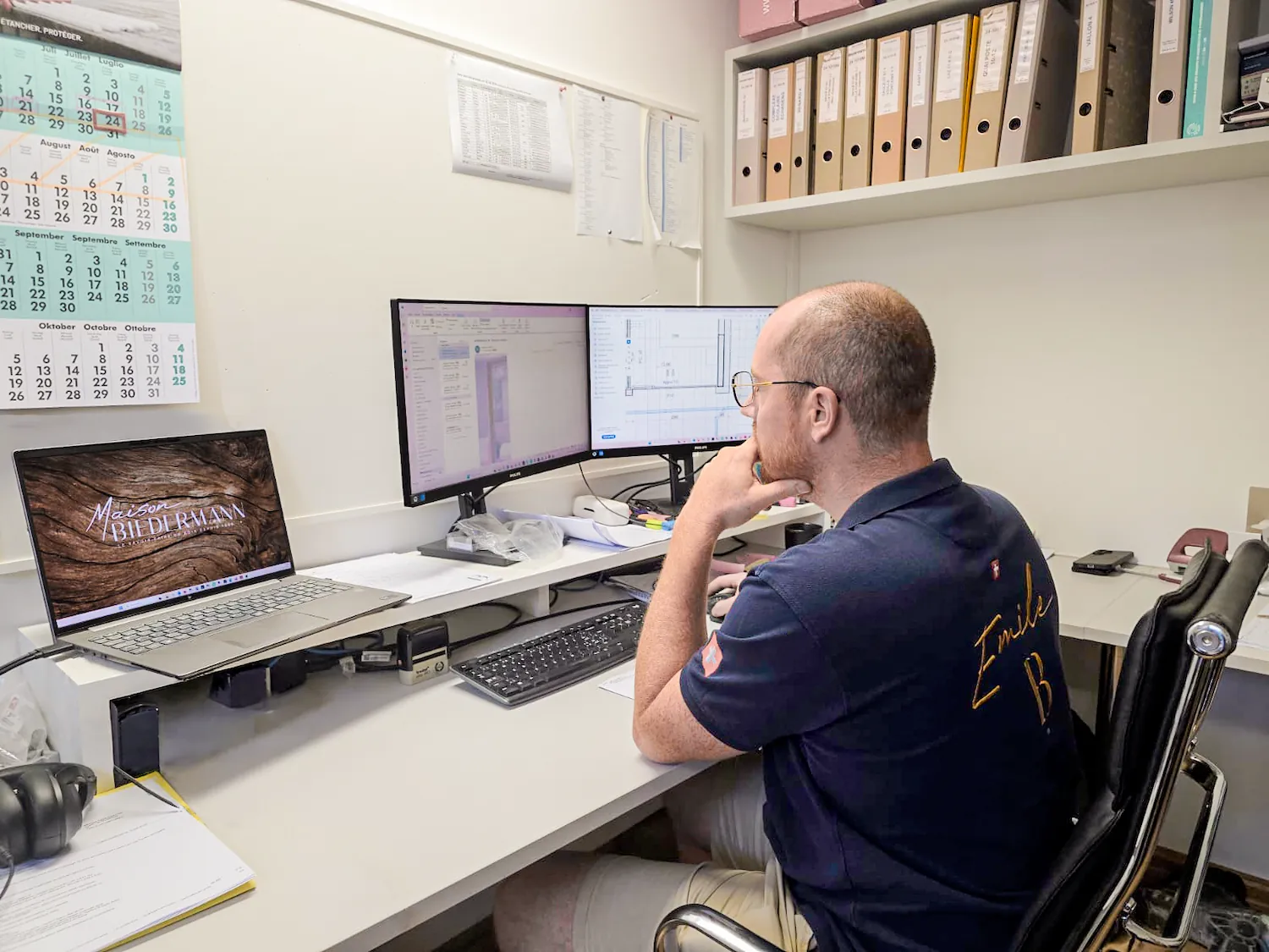 Collaborateur d'Emile B au bureau, polo floqué « Emile B », consultant des plans techniques sur deux écrans et un ordinateur portable.