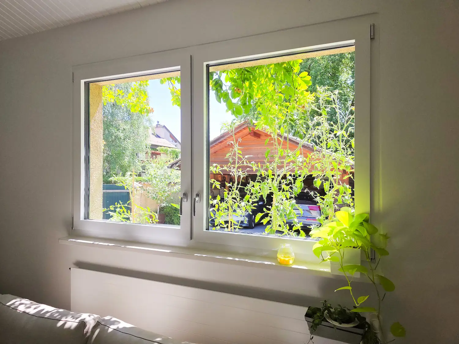 Fenêtre à deux vantaux blanche vue de l'intérieur, ouvrant sur un jardin arboré avec un chalet en bois, plante verte posée sur l'appui de fenêtre.