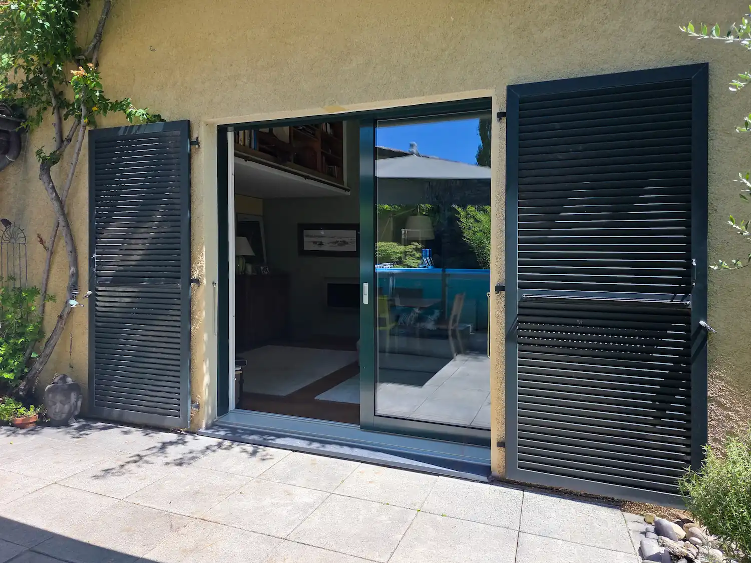 Porte-fenêtre coulissante vitrée à cadre gris anthracite encadrée de deux volets battants persiennés anthracite, sur une façade crépie jaune ocre, donnant sur un salon et une piscine.